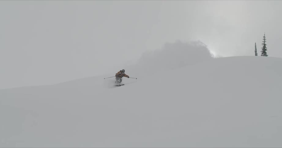 A skier kicking up snow as they ski down hills on a cloudy day