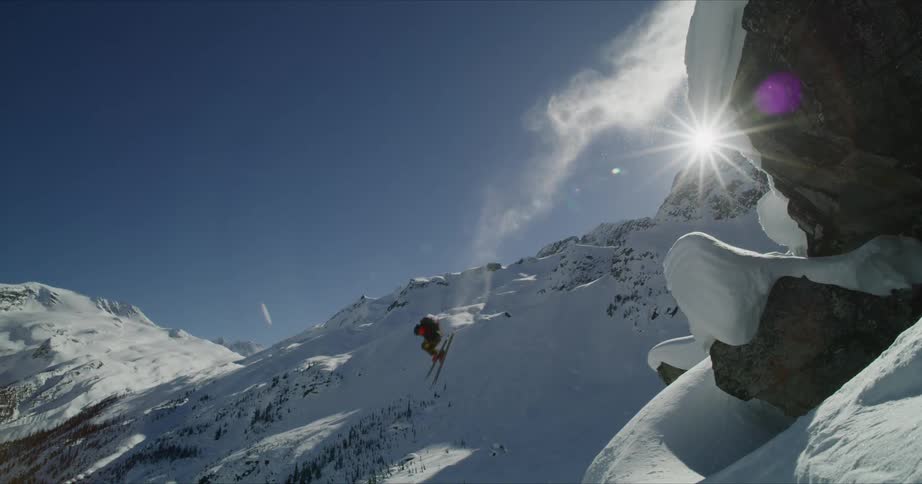 A skier jumps off a mountain ridge onto a slope on a sunny blue sky day