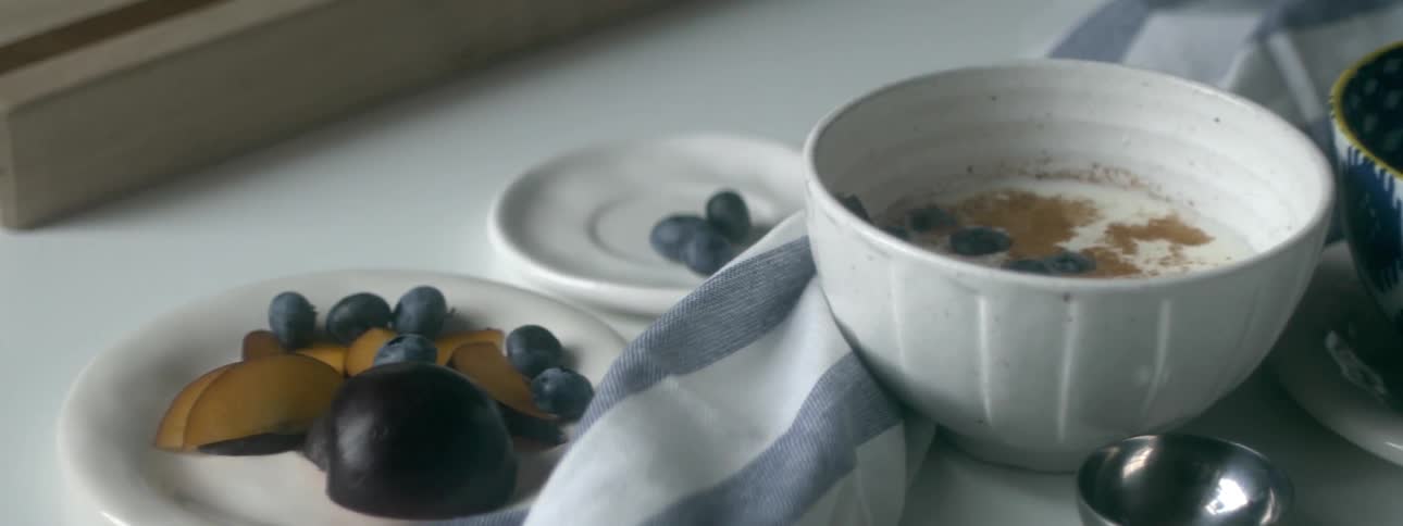 Plums and blueberries are placed on a saucer next to a bowl of cereal on a dining table