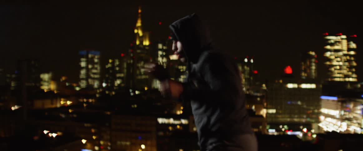 Man wearing a hoodie boxing on a rooftop at night against the city lights