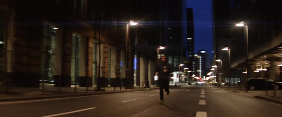 A man is running on a downtown street at night with a glimpse of an ink blue sky in the distance seen between two buildings