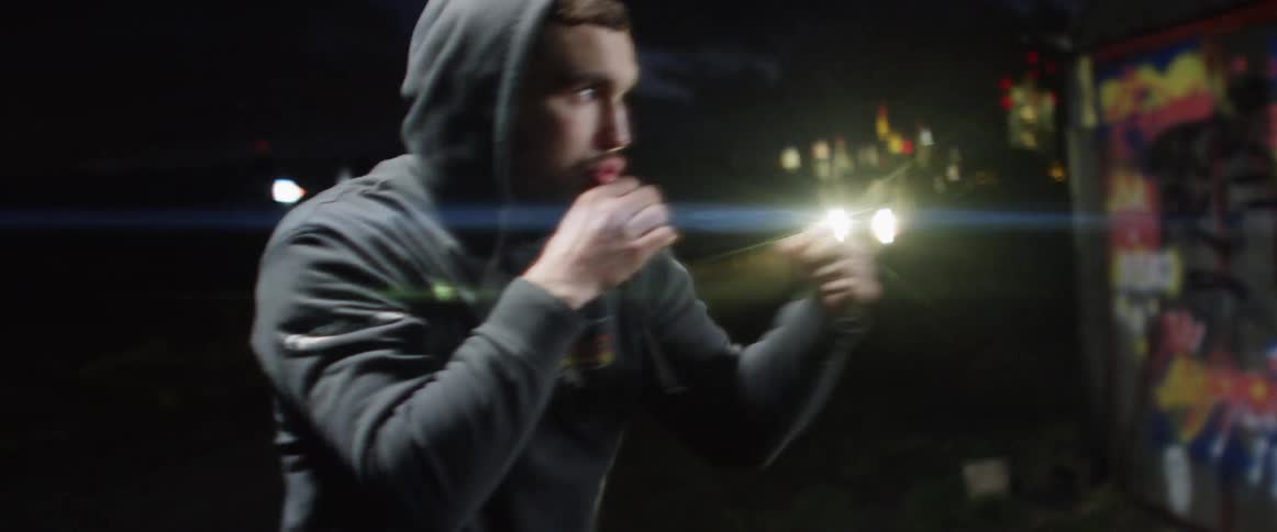 A man in a hoodie shadow boxes near a graffiti covered fence at night