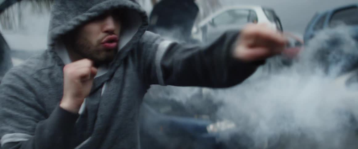A man boxes in a car junkyard as smoke from a car exhaust surrounds him