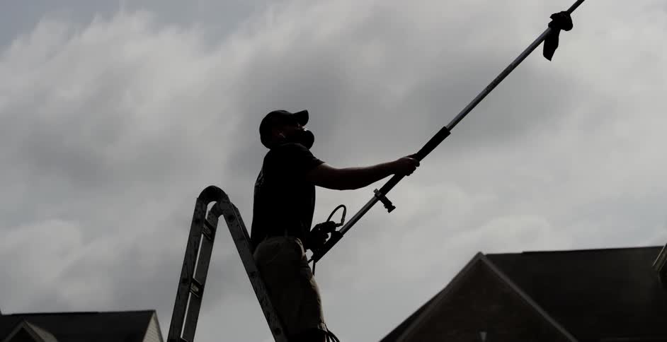 A man stands on a ladder wearing a mask as he controls a long rod power washer to clean a house