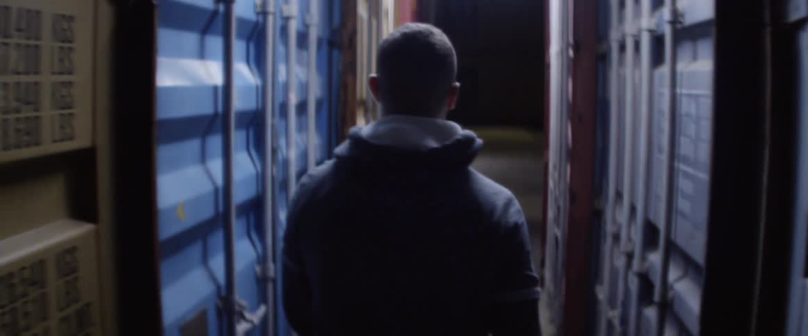 A man in a hoodie walking between two rows of shipping containers