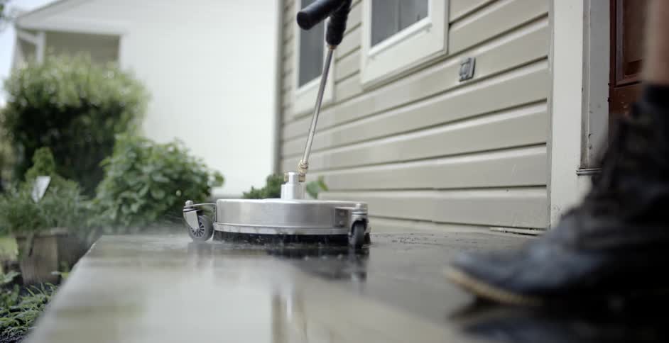 A person wearing boots walks backwards as he pressure washes concrete outside a house