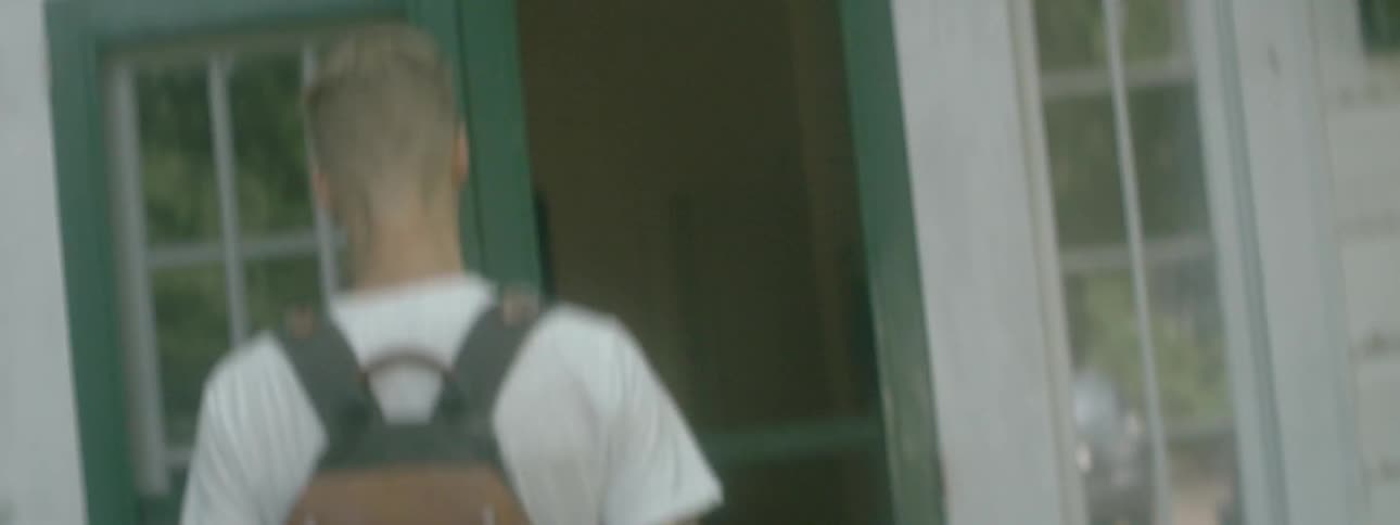 A man carrying a skateboard and wearing a backpack while walking up a sidewalk and opening a screen door