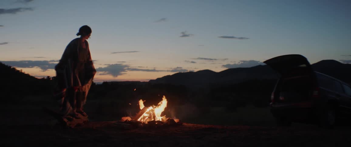 Woman wearing poncho standing in front of campfire at sunset