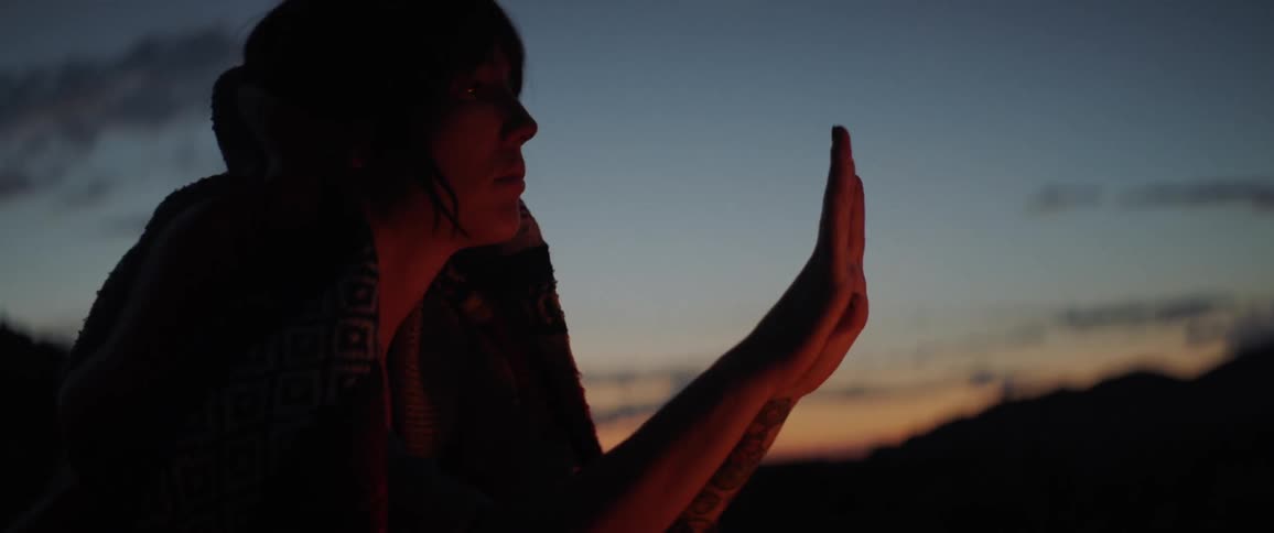A young woman sits outside warming her hands by a fire just after sunset