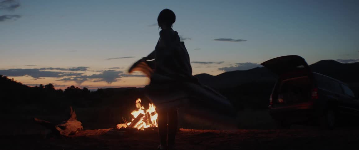 A woman stands in front of a campfire at dusk while playing with a shawl