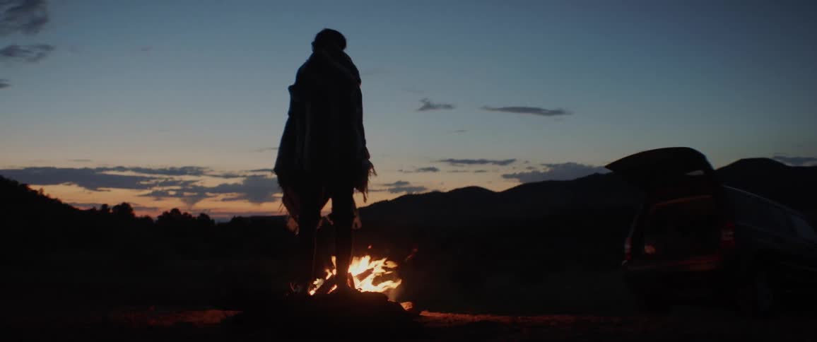 Person standing by campfire angrily wrapping blanket around them at sunset