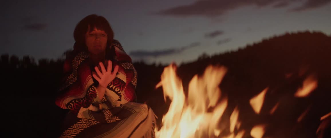 Woman covered with blanket sits down next to the campfire at dusk