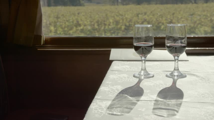 Two glasses of red wine sit atop a table next to the window of a moving train that is passing a large field