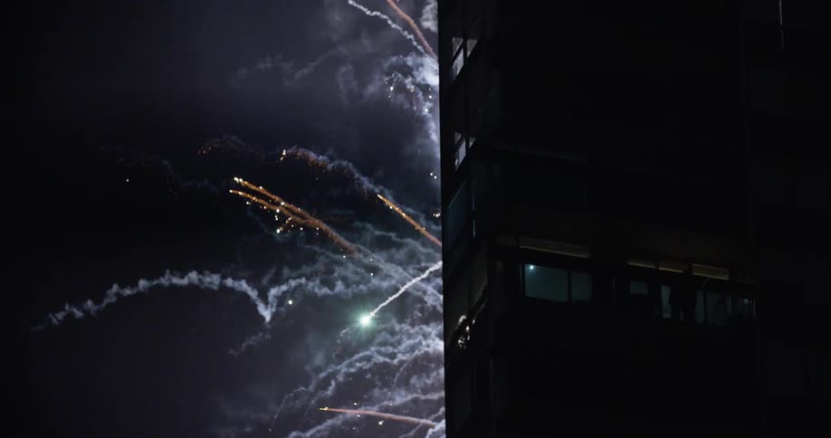 People on building balconies stand and watch fireworks shooting into the sky