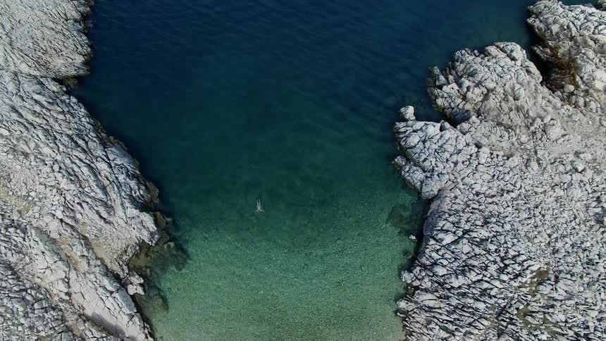 A lone person swims in a large body of water surrounded by rocky shores and a shallow rocky beach