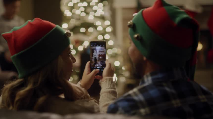A woman takes a picture of a christmas tree while wearing an elf hat and sitting next to a man