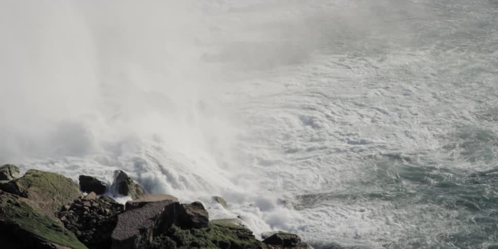 Water churns and sprays as it flows over waterfall past rocky shore