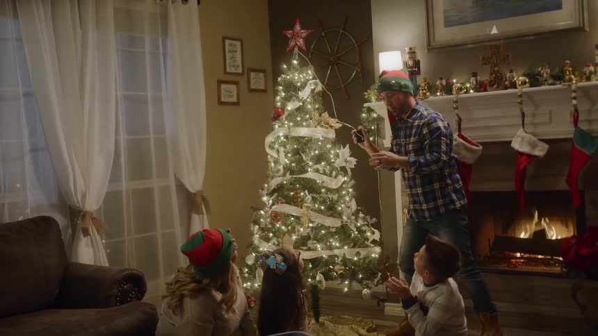 Father placing star on top of christmas tree and turning lights on as family claps