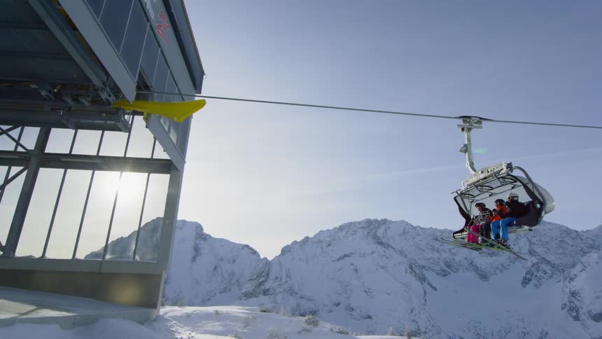 Three people ride on a ski lift and prepare to exit it at the top of a snowy mountain
