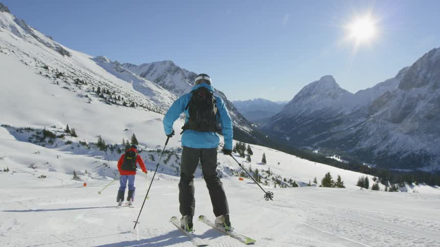 People ski down slight slope surrounded by snow covered mountains