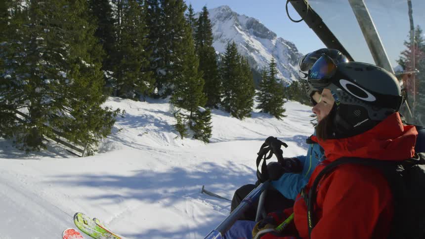 Two white men decked out in skiing gear ride a ski lift up a slope