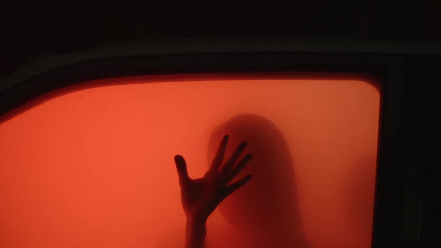 A person, with their hand on the window, sits in a car filled with fog lit by a reddish light