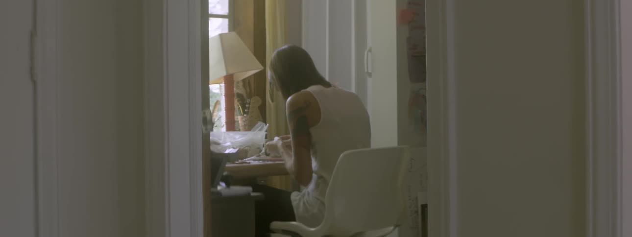 A tattooed woman sits in front of a window string jewelry