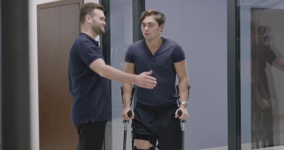 A patient with crutches and knee brace meets a therapist and walks down a hall
