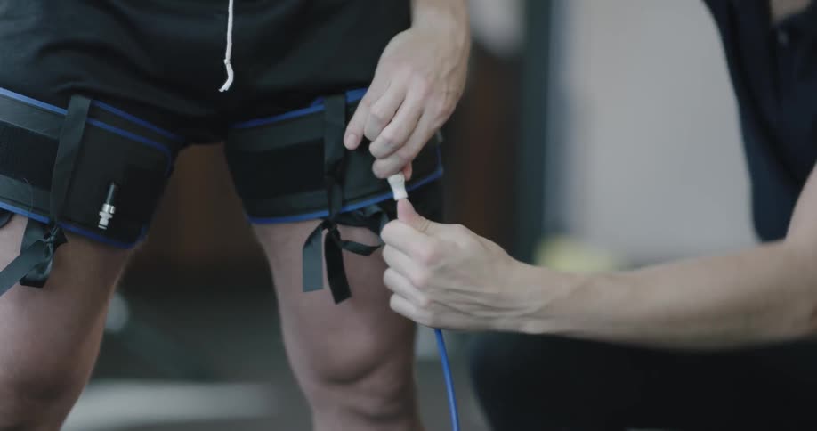 A man in shorts is standing with a cuff wrapped around each of this thighs as a sports trainer connects a cable to one of them