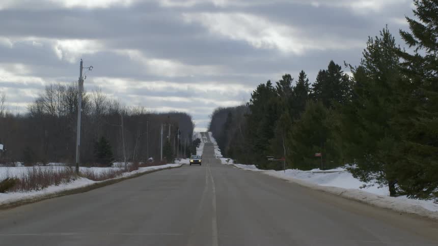 View of a car driving down a hilly, tree-lined road on a cloudy winter day