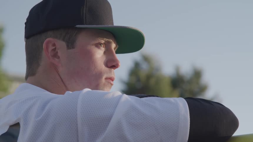 Baseball player wearing a cap rests his elbows on the banister and looks ahead on a sunny day