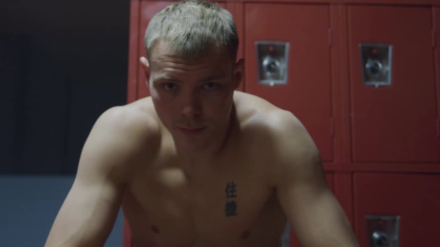Male boxer with a shirt sits on a bench in the locker room swaying back and forth with his boxing gloves on