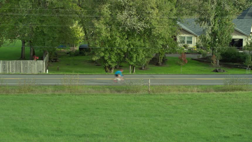 A bicyclist speeds along a quiet neighborhood street lined by a grassy field
