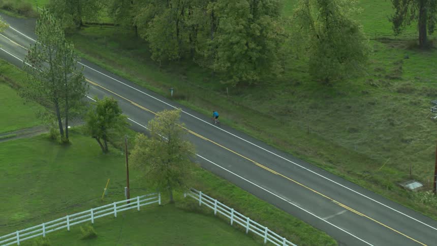 An aerial view of a bicyclist riding on a rural road near green trees and grass