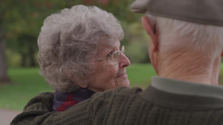 A woman looks at her husband who has his arm around her while they sit on a park bench