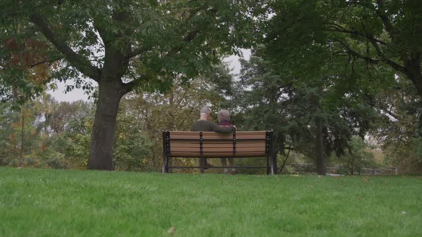 A husband puts his arm around his wifes shoulder as they sit on a bench in the park