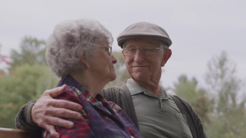 Senior man and woman sitting on the bench hold each other in the public park