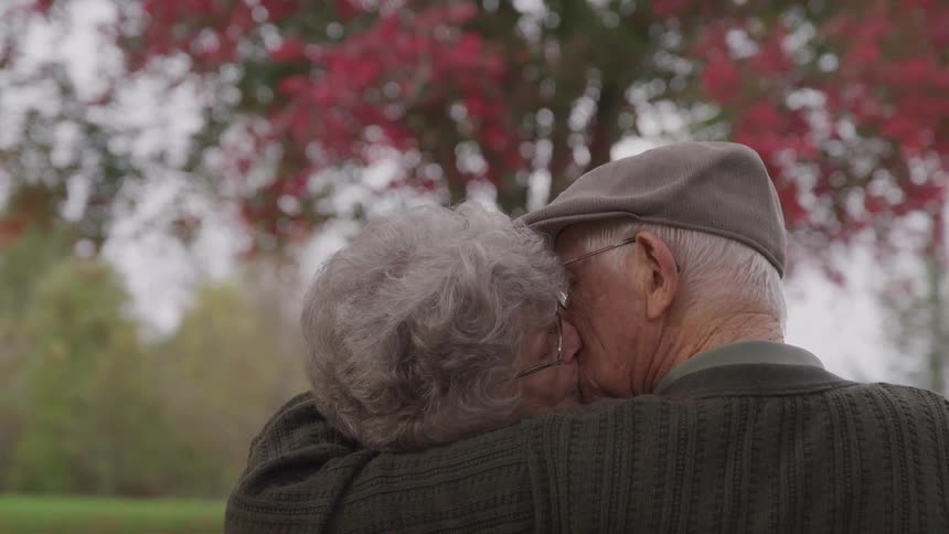 Senior man and woman sitting on the park bench kiss each other in the public park