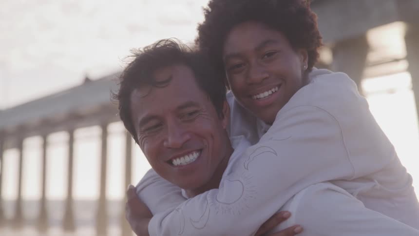 Woman piggy back rides on man near pier on beach at sunset