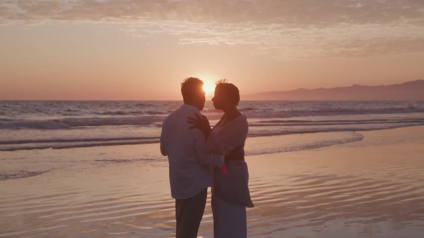 Husband and wife are holding each other while the sun sets by the ocean coast