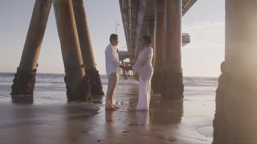 Couple hold hands under pier on the beach at sunset next to the ocean