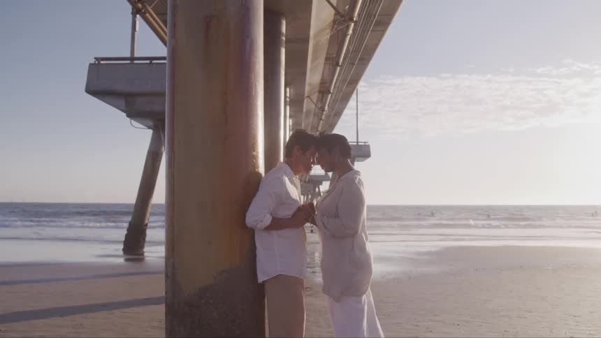 A husband and wife hold hands as they lean against a pole on the beach