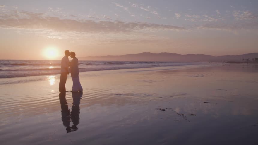 Asian man and black woman stands on the sand beach and hold each other at sunset next to the ocean