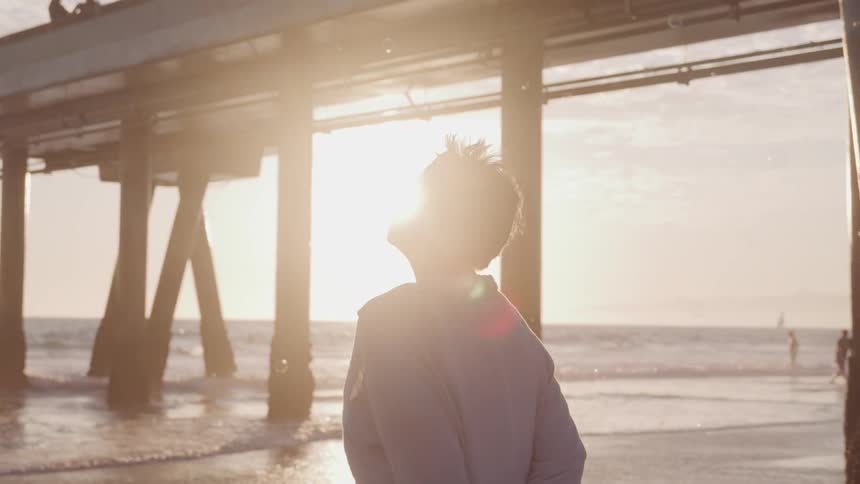 A woman stands on the beach by a large pier looking up to the sky