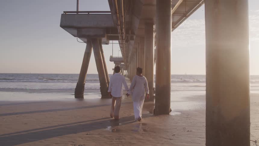 A man and woman hold hands as they walk on the beach under a pier
