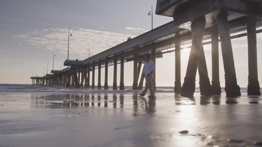 Man walks under pier on beach next to the ocean at sunset