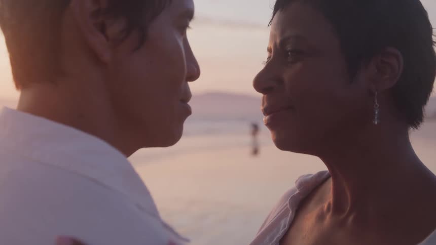 A man and woman stare lovingly into each others eyes while standing on a beach during sunset