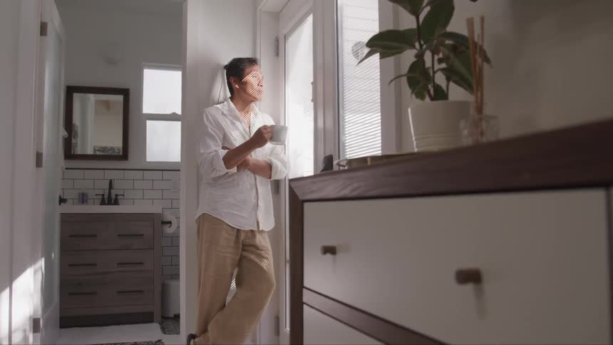 A man leans against the wall drinking coffee and looking out the door on a sunny day