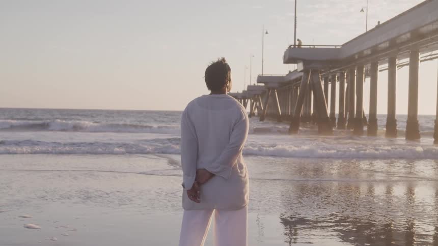A woman stands on the beach near the pier pylons looking out at the ocean
