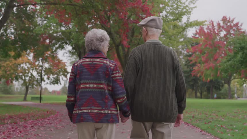 An elderly couple walks hand in hand on a park path in autumn